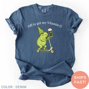 Op de afbeelding: Denim blauw t-shirt met een humoristische illustratie van een groene kikker met een tovenaars hoed, die een biertje vasthoudt en op een scooter rijdt. De tekst erboven luidt "Off to get my Vitamin G".
