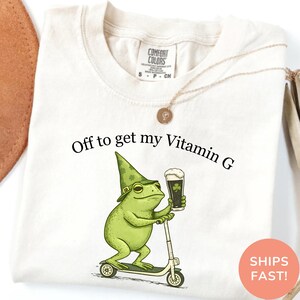 Op de afbeelding: Cr&egrave;mekleurig T-shirt met de tekst "Off to get my Vitamin G." Een groene kikker met een kabouterhoed en een pint stout bier met een klaverblad rijdt op een scooter. Het shirt heeft een Comfort Colors label.