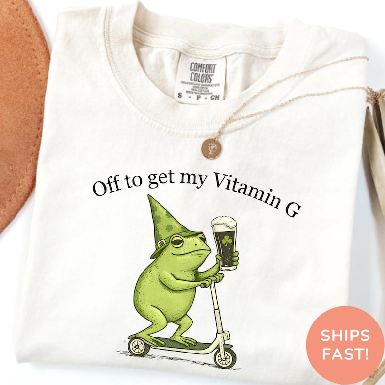 Op de afbeelding: Cr&egrave;mekleurig T-shirt met de tekst "Off to get my Vitamin G." Een groene kikker met een kabouterhoed en een pint stout bier met een klaverblad rijdt op een scooter. Het shirt heeft een Comfort Colors label.