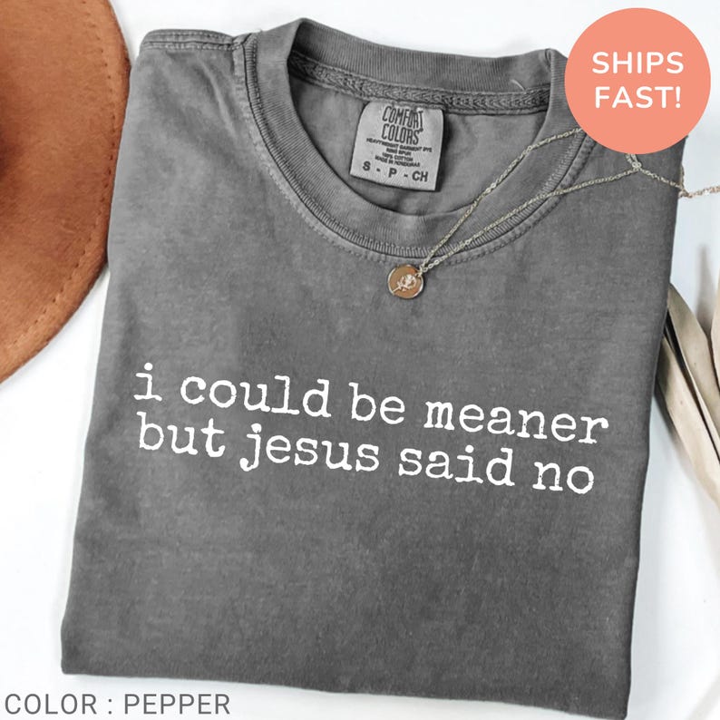 May include: A gray t-shirt with the text "i could be meaner but jesus said no" in white. The shirt has a crew neck and short sleeves. A gold necklace with a pendant is on top of the shirt.