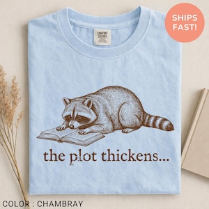 May include: Pale blue t-shirt featuring a raccoon illustration reading a book. The text "the plot thickens..." is printed below the raccoon. The shirt is labelled "Comfort Colors" and the colour is "Chambray".