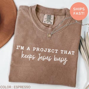 May include: Espresso-coloured t-shirt with the text "I'M A PROJECT THAT keeps Jesus busy" in white. The shirt has a crew neck and a small tag near the neckline. A silver necklace with a pendant is draped across the shirt.