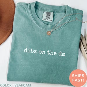 May include: Seafoam green t-shirt with the phrase "dibs on the dm" in white lettering. The shirt has a crew neck and short sleeves. A silver necklace with a small pendant rests on the shirt. The shirt is labeled "Comfort Colors".