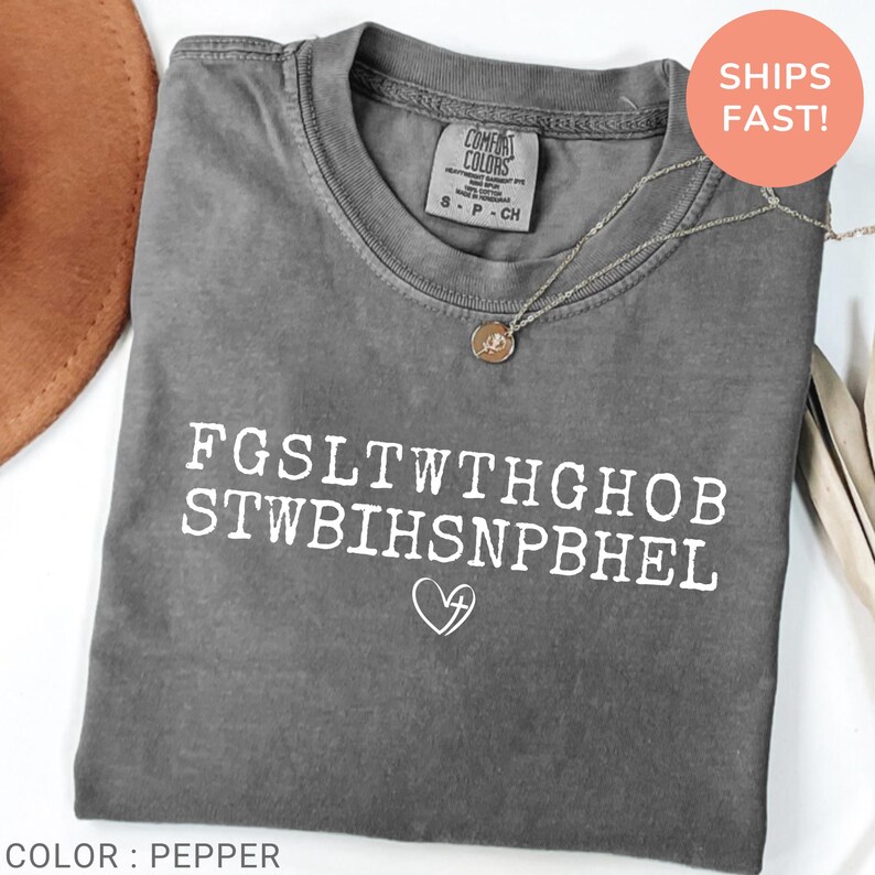 Pu&ograve; includere: Una maglietta grigia Comfort Colors con testo bianco e un simbolo a forma di cuore. La maglietta ha il testo "FGSLTWTHGHOB STWBIHSNPBHEL" stampato sul davanti. Una collana d'argento con un ciondolo &egrave; drappeggiata sulla maglietta. La maglietta &egrave; etichettata "Color: Pepper".