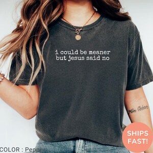 May include: Pepper-colored t-shirt with the text "i could be meaner but jesus said no" in white. The shirt is a dark gray color and has a crew neck. The image also includes a necklace and a tattoo on the arm.
