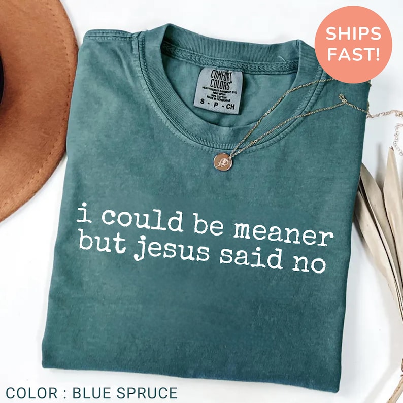 May include: Blue spruce t-shirt with the text "i could be meaner but jesus said no" in white. The shirt has a crew neck and a small tag near the collar. A gold necklace with a pendant is draped across the shirt.