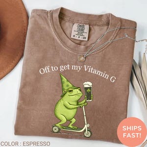 Op de afbeelding: Espresso-kleurig t-shirt met de tekst "Off to get my Vitamin G". Een groene kikker met een hoed en een bierglas met een klavertje vier rijdt op een scooter. Het shirt heeft een Comfort Colors-label.