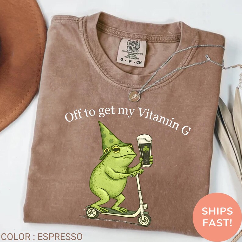 Op de afbeelding: Espresso-kleurig t-shirt met de tekst "Off to get my Vitamin G". Een groene kikker met een hoed en een bierglas met een klavertje vier rijdt op een scooter. Het shirt heeft een Comfort Colors-label.