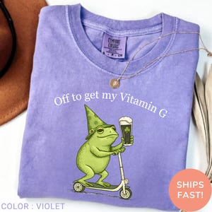 Op de afbeelding: Violet Comfort Colors T-shirt met een afbeelding van een groene kikker met een hoed en een biertje op een scooter. De tekst "Off to get my Vitamin G" staat erboven. Ronde hals.
