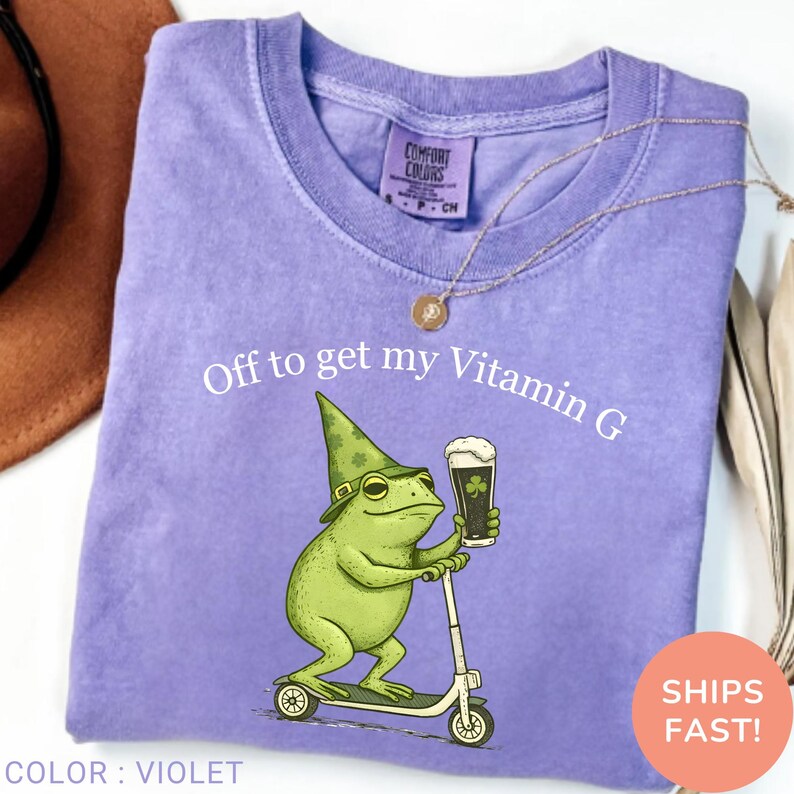 Op de afbeelding: Violet Comfort Colors T-shirt met een afbeelding van een groene kikker met een hoed en een biertje op een scooter. De tekst "Off to get my Vitamin G" staat erboven. Ronde hals.
