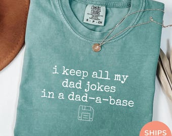 May include: Seafoam green t-shirt with the phrase "i keep all my dad jokes in a dad-a-base" in white text. A floppy disk graphic is below the text. A silver necklace with a pendant is on the shirt.