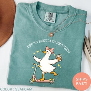 May include: Seafoam green Comfort Colors t-shirt with the phrase "OFF TO REGULATE EMOTIONS" and a cartoon duck riding a pink scooter, holding a coffee cup. The shirt has a crew neck and short sleeves.