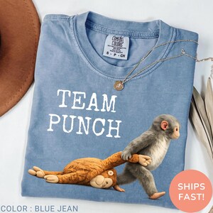 May include: Blue jean-colored t-shirt with the text "TEAM PUNCH" in white. The shirt features a cartoon monkey pulling another monkey. A gold necklace is draped on the shirt.