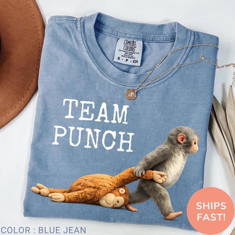 May include: Blue jean-colored t-shirt with the text "TEAM PUNCH" in white. The shirt features a cartoon monkey pulling another monkey. A gold necklace is draped on the shirt.