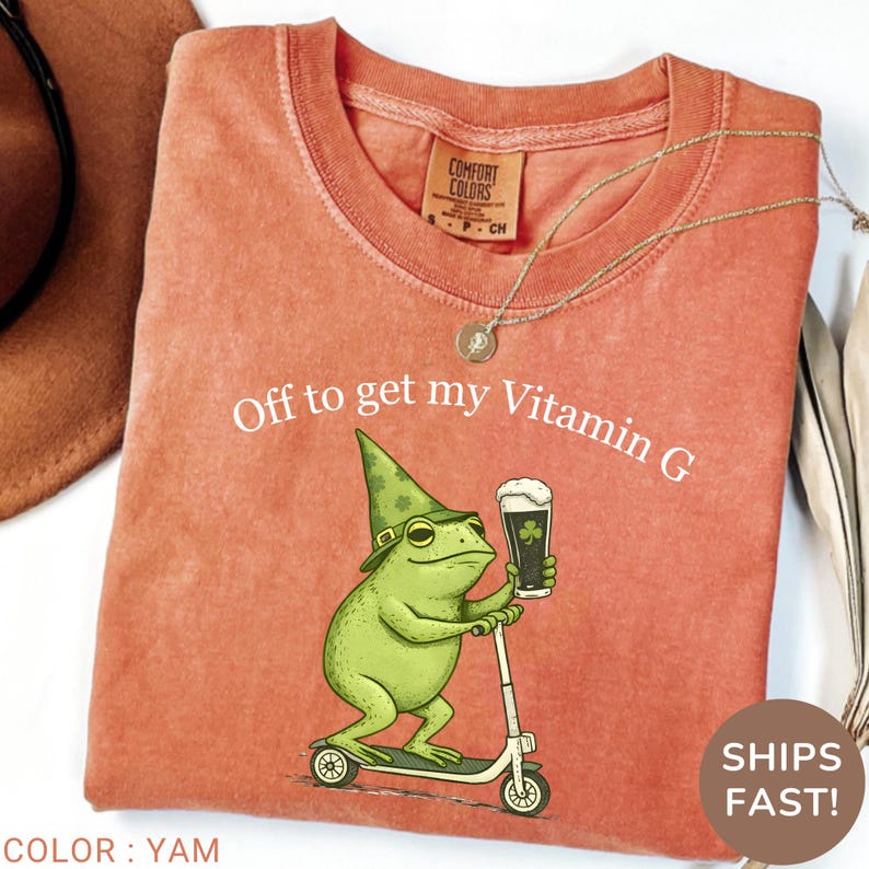 Op de afbeelding: Een yamkleurig T-shirt met de tekst "Off to get my Vitamin G". De afbeelding toont een groene kikker met een hoed die op een scooter rijdt en een biertje vasthoudt. Het shirt heeft een Comfort Colors-label.