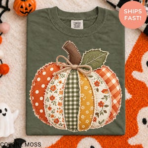 Peut inclure: Un t-shirt vert olive arborant un grand motif central de citrouille composé de divers empiècements en tissu. Le design de la citrouille inclut des pois orange, des motifs floraux, du vichy vert et du tartan orange, avec un nœud en jute.