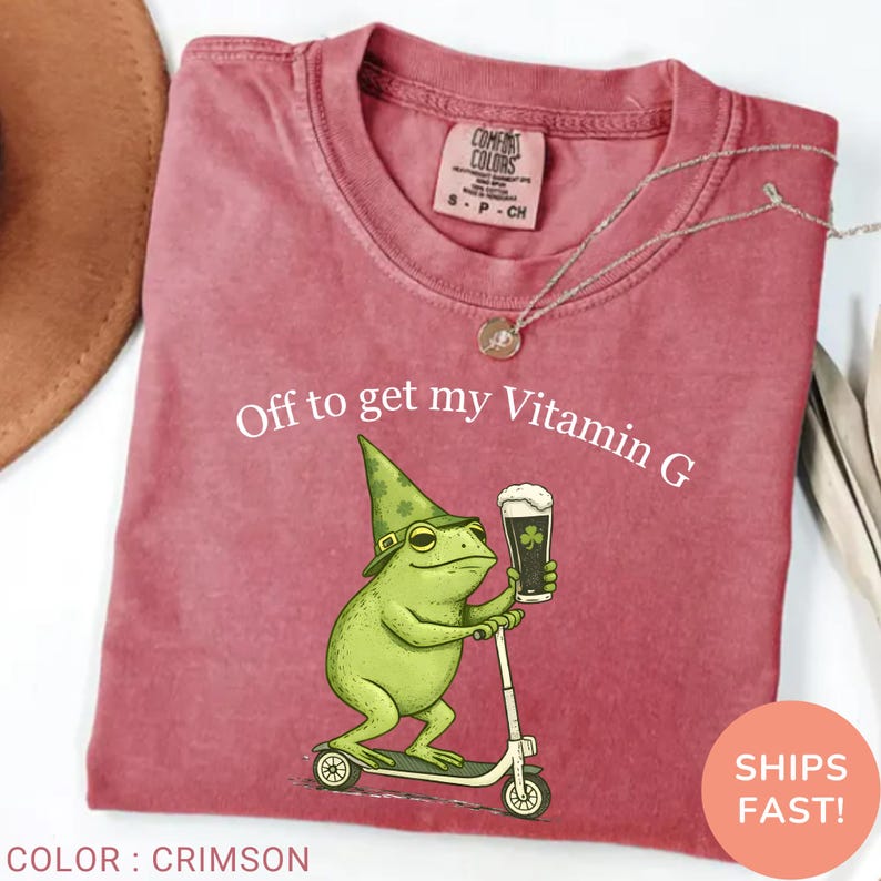 Op de afbeelding: Karmizijnrood T-shirt met een afbeelding van een groene kikker met een hoed, die een biertje vasthoudt en op een scooter rijdt. De tekst "Off to get my Vitamin G" staat boven de kikker.