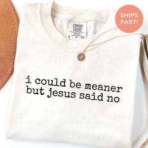 May include: Cream-colored t-shirt with the text "i could be meaner but jesus said no" in black. The shirt has a crew neck and short sleeves. A gold necklace with a pendant is draped across the shirt.