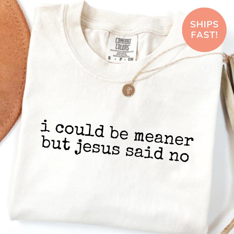 May include: Cream-colored t-shirt with the text "i could be meaner but jesus said no" in black. The shirt has a crew neck and short sleeves. A gold necklace with a pendant is draped across the shirt.