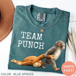 May include: Blue spruce Comfort Colors t-shirt with the text "TEAM PUNCH" in white. A cartoon monkey pulls another monkey. A gold necklace is draped on the shirt. The words "SHIPS FAST!" are in a red circle.