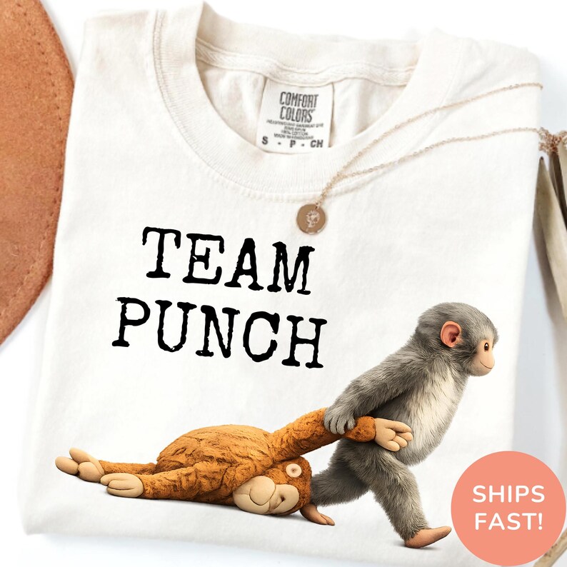 May include: Cream-colored t-shirt with the text "TEAM PUNCH" in black. A cartoon monkey is pulling another cartoon monkey. A gold necklace is draped on the shirt. A round sticker with the text "SHIPS FAST!" is in the bottom right corner.