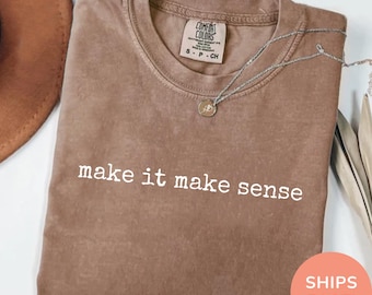 May include: Espresso-coloured t-shirt with the phrase "make it make sense" in white block letters. The shirt has a crew neck and short sleeves. A silver necklace with a small pendant is draped across the shirt.