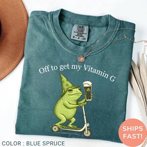 Op de afbeelding: Blauwspar T-shirt met de tekst "Off to get my Vitamin G". De afbeelding toont een groene kikker met een hoed en bier op een scooter. Het shirt is van het merk Comfort Colors.