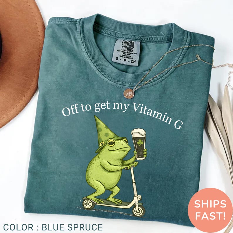 Op de afbeelding: Blauwspar T-shirt met de tekst "Off to get my Vitamin G". De afbeelding toont een groene kikker met een hoed en bier op een scooter. Het shirt is van het merk Comfort Colors.