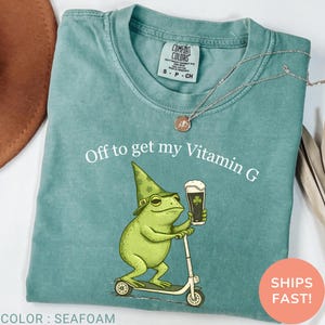Op de afbeelding: Zeegroen t-shirt met de tekst "Off to get my Vitamin G." De afbeelding toont een groene kikker met een hoed en een biertje, op een scooter. Het shirt heeft een Comfort Colors label.