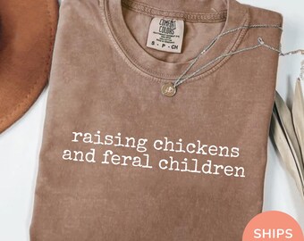 May include: Espresso-colored t-shirt with the text "raising chickens and feral children" in white. The shirt has a crew neck and short sleeves. A silver necklace with a pendant is draped across the shirt.