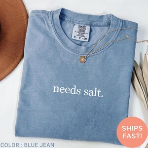 May include: A blue Comfort Colors t-shirt with the phrase "needs salt" printed on it. A gold necklace with a pendant is on the shirt. The shirt is folded, and a brown hat is partially visible in the background.