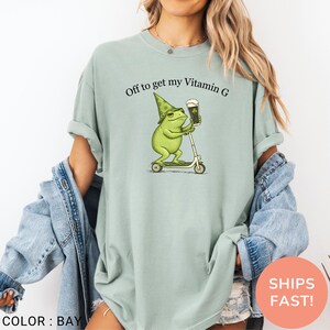 Op de afbeelding: Lichtgroen T-shirt met de tekst "Off to get my Vitamin G". Het shirt heeft een cartoon kikker met een groene hoed die een biertje vasthoudt en op een scooter rijdt. Het shirt is effen van kleur.