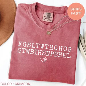 Pu&ograve; includere: T-shirt color cremisi con testo bianco e un cuore con grafica a croce. La maglietta ha uno scollo tondo e una piccola etichetta sullo scollo. Una collana color oro &egrave; drappeggiata sulla maglietta.