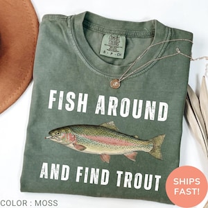 May include: A moss green t-shirt featuring the text "FISH AROUND AND FIND TROUT" and a trout illustration. The shirt has a "COMFORT COLORS" tag. A necklace and a hat are also in the image. The shirt is a relaxed fit.
