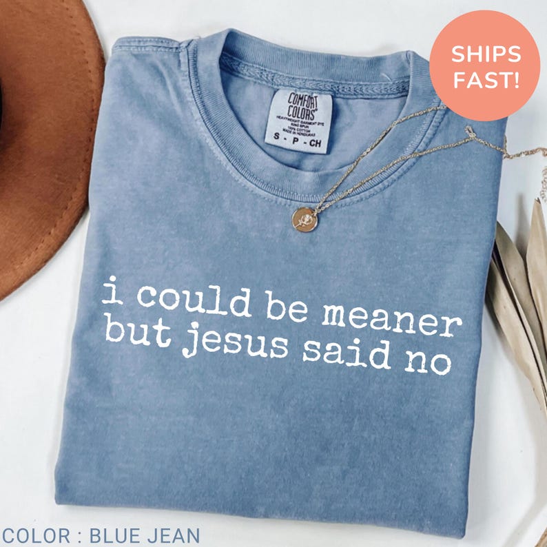 May include: Blue jean colored t-shirt with the text "i could be meaner but jesus said no" in white. A gold necklace with a pendant rests on the shirt. The shirt has a Comfort Colors tag.