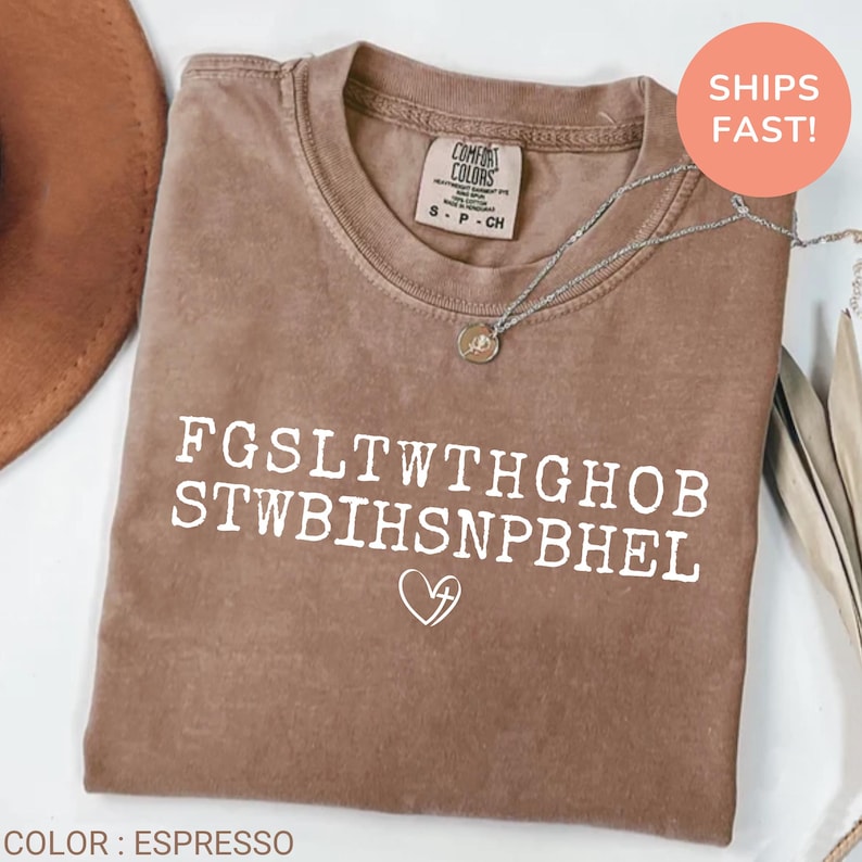 Pu&ograve; includere: T-shirt Comfort Colors color espresso con testo bianco e simbolo a cuore. La maglietta ha girocollo e maniche corte. Una collana d'argento con un ciondolo &egrave; drappeggiata sulla maglietta.