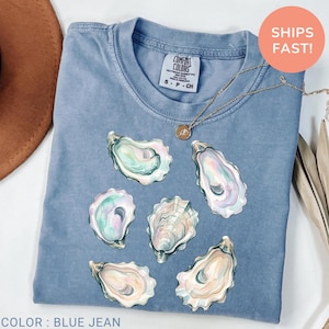 May include: Blue jean-colored t-shirt featuring watercolor oyster shell graphics. The shirt has a crew neck and short sleeves. A gold necklace with a pendant is draped across the shirt. The tag reads "Comfort Colors". The text "Color: Blue Jean" is at the bottom.