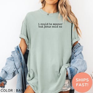 May include: A light green t-shirt with the text "i could be meaner but jesus said no" in black. The shirt has short sleeves and a crew neck. The color is "bay".