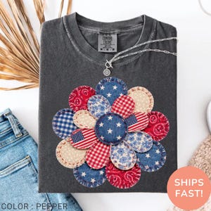 May include: Dark gray Comfort Colors t-shirt with a floral design made of red, white, and blue fabric patches. The patches feature stars, stripes, and floral patterns. The text "COLOR: PEPPER" is at the bottom.