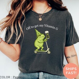 Op de afbeelding: Donkergrijs t-shirt met de tekst "Off to get my Vitamin G". Een groene kikker met een puntmuts en een bierpul met een klavertje rijdt op een scooter. Het shirt heeft de kleur peper.