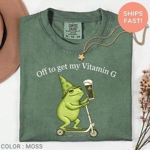 Op de afbeelding: Mosgroen t-shirt met de tekst "Off to get my Vitamin G". Het shirt heeft een cartoon kikker met een groene hoed, die een biertje vasthoudt en op een scooter rijdt. Een gouden sterrenketting is zichtbaar.