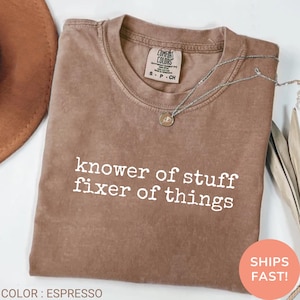 May include: Espresso-colored t-shirt with the text "knower of stuff fixer of things" in white. The shirt has a crew neck and a Comfort Colors tag. A silver necklace with a pendant is draped across the shirt.