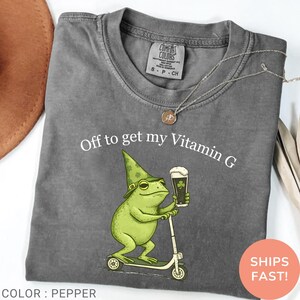Op de afbeelding: Pepperkleurig t-shirt met een afbeelding van een groene kikker op een scooter, met een hoed en een biertje. De tekst "Off to get my Vitamin G" staat boven de kikker. Een ketting en een hoed zijn zichtbaar.