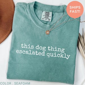 May include: A seafoam green short-sleeve t-shirt with a crew neckline. The shirt features the white text "this dog thing escalated quickly" printed across the chest. A silver-toned necklace with a small circular pendant rests on the neckline.