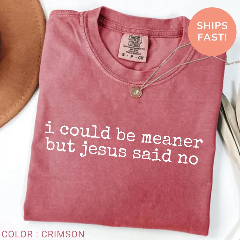 May include: Crimson-colored t-shirt with the text "i could be meaner but jesus said no" in white. The shirt has a crew neck and a small tag at the neckline. A gold-colored necklace with a pendant is draped over the shirt.