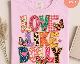 May include: A light pink t-shirt featuring the text LOVE LIKE DOLLY in large, multi-patterned letters. The design includes floral, gingham, cow print, leopard print, and star patterns, accented with rhinestones and three colorful butterflies.