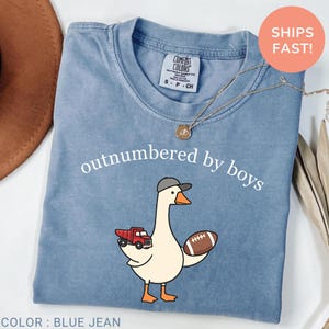 Peut inclure: T-shirt bleu jean avec l'inscription "outnumbered by boys". Le t-shirt présente un dessin d'oie de dessin animé portant une casquette de baseball, tenant un camion rouge et un ballon de football. Un collier en or est drapé sur le t-shirt.