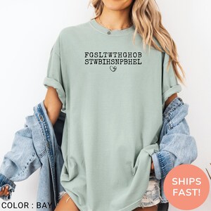 Pu&ograve; includere: T-shirt verde chiaro con testo nero e simbolo a cuore. Il testo recita "FGSLTWTHGHOB STWBIHSNPBHEL". La maglietta &egrave; di colore tinta unita e ha maniche corte.