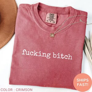 May include: Crimson t-shirt with the words "fucking bitch" in white. The shirt has a crew neck and a small tag on the upper chest. A silver necklace with a pendant is draped on the shirt. The colour is described as crimson.