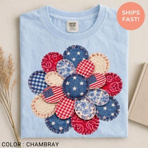 May include: Light blue t-shirt with a floral design made of various fabric patterns, including American flags, stars, and gingham. The shirt is labeled "Comfort Colors" and the color is "Chambray". A round sticker says "Ships Fast!"